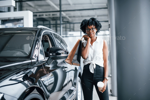 Young african american woman in glasses stands in car salon near vehicle with package in hands