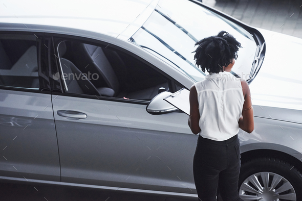 Young african american woman in glasses and with notepad stands in the car salon near vehicle