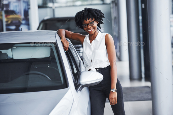 Young african american woman in glasses stands in the car salon near vehicle