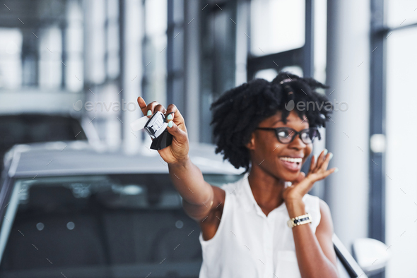 Young african american woman in glasses stands in the car salon near vehicle with keys in hands