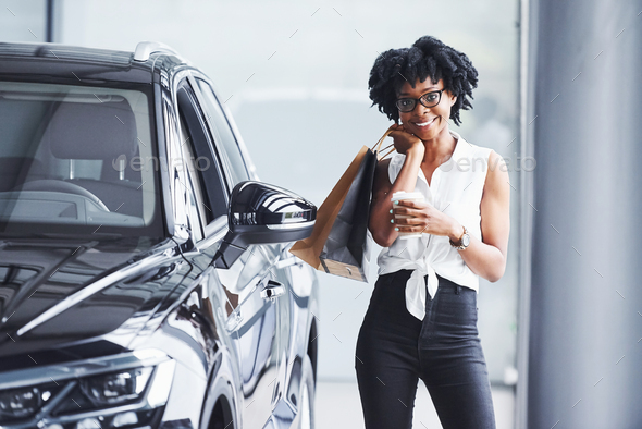 Young african american woman in glasses stands in car salon near vehicle with package in hands
