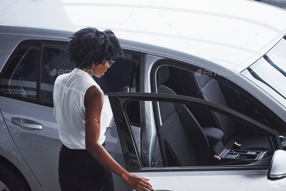 Young african american woman in glasses and with notepad stands in the car salon near vehicle