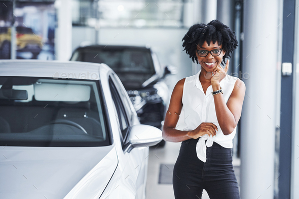 Young african american woman in glasses stands in the car salon near vehicle