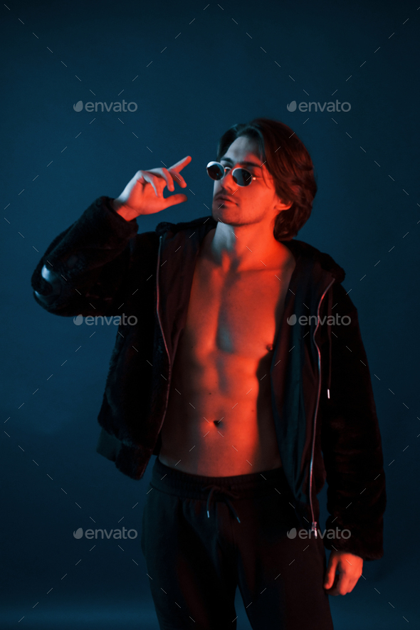 Beautiful man in glasses and black clothes is in the studio with blue neon lighting