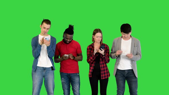 Young Friends Stand in Front of Camera Focused on Their Gadgets on a Green Screen Chroma Key