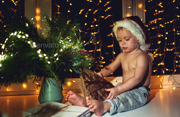 A beautiful little boy in a Santa hat sits by the window at Christmas time