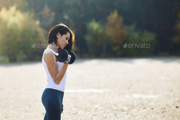 A brunette girl in a hoodie came outside to do fitness with a Mat and dumbbells.