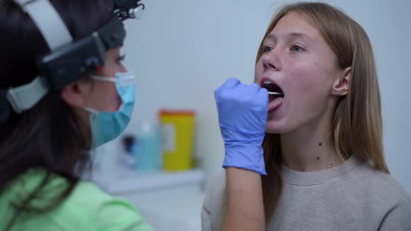 Portrait of Teenage Caucasian Girl Opening Mouth for Medical Examination As Blurred Doctor Checking alt