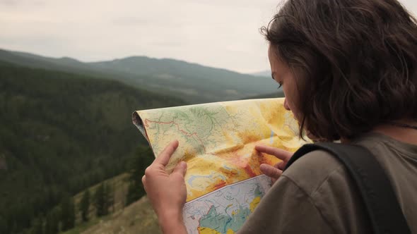A young girl on a trip stands with a paper map against the background of mountains and a lake.