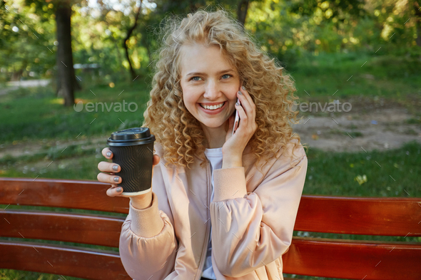 Outdoor photo of young beautiful girl blonde sits on a park bench, drinking coffee, speaking
