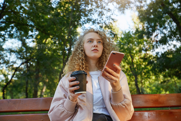 A young beautiful girl blonde sits on a park bench, drinking coffee, looks on a companion, speaking