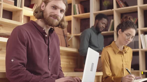 Smiling Young Male Hipster Working on Laptop in Book Cafe alt