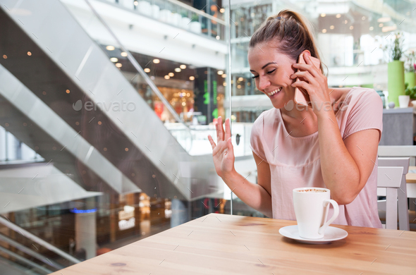 Girl before meeting with friend talking on phone and waving in cafe at shopping mall