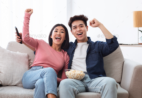 Excited Chinese Couple Watching TV Shaking Fists In Living Room