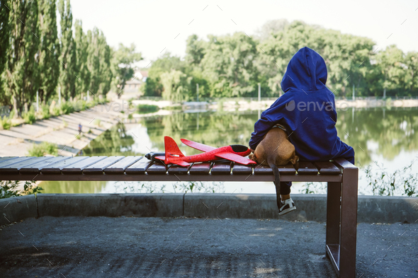 Sad kid sitting alone with dog and toy airplane on bench by the river bank. Diagnosing Early-Onset - Stock Photo - Images