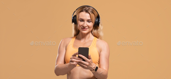 Smiling muscular middle aged woman in sportswear with headphones listen song on smartphone
