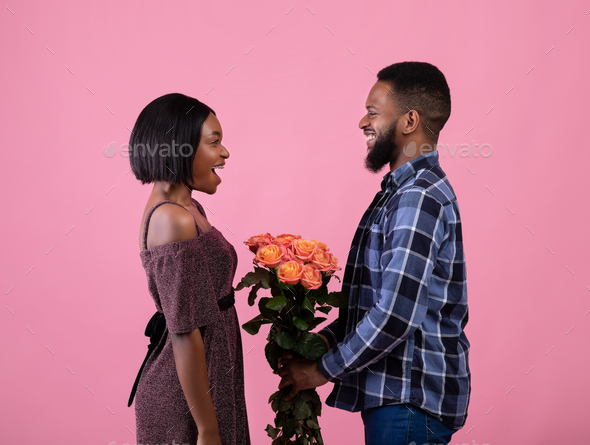 Side view of positive black man presenting flowers to his girlfriend over pink studio background