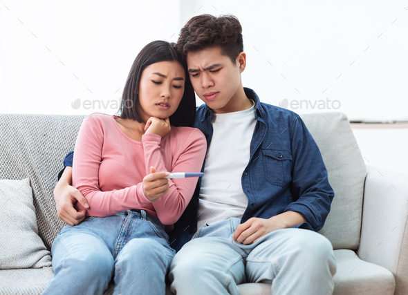Frustrated Asian Couple Holding Negative Pregnancy Test Sitting At Home