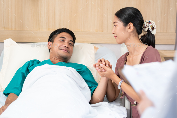 Asian woman visiting male patient on bed at hospital ward - Stock Photo - Images