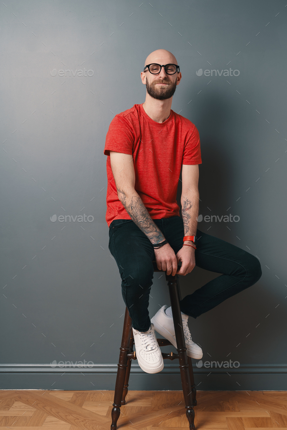 Young smiling man with beard and glasses sitting on a tall chair on gray studio  background Stock Photo by diignat