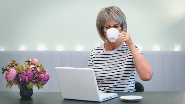 An Elderly Beautiful Woman Works at Home at a Laptop in the Home Office