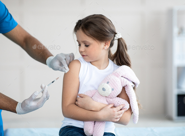 Doctor doing injection of coronavirus vaccine to little girl with toy bunny at clinic. Medicine and