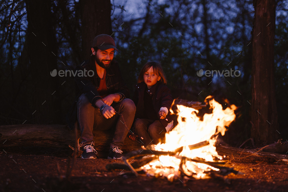 The father and his little son sitting on the logs in the forest in front of a fire and roasting