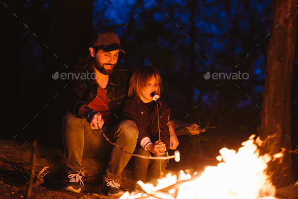 Happy father and his little son sitting on the logs in the forest in front of a fire and eating