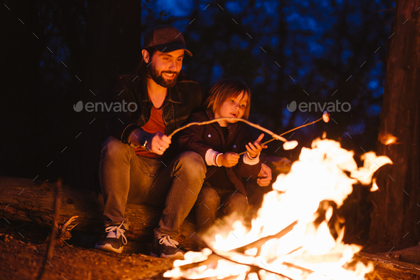 Happy father and his son sitting on the logs in the forest in front of a fire and roasting