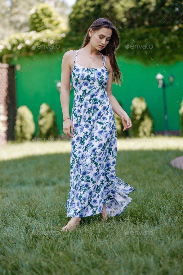 Brunette girl dressed in a stylish summer sundress walks barefoot on the  lawn in the garden on a Stock Photo by leikapro