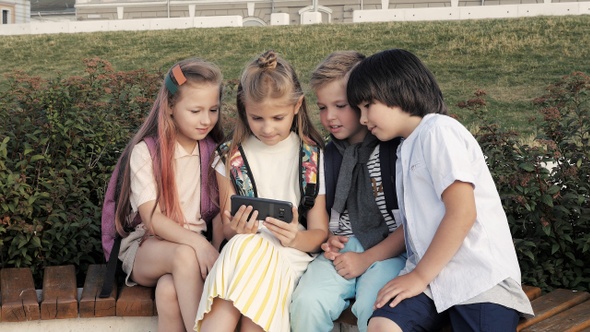School friends watching video on the phone sitting on a bench. alt