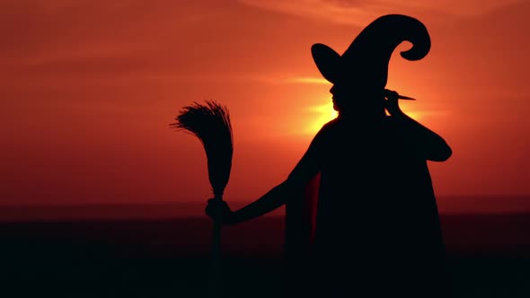 Girl in Fancy Dress Witch Holds a Broom Looking on Sunrise alt