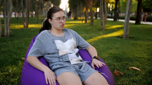 A Teenage Girl with Glasses in a City Park is Sitting on a Large Soft Pillow alt