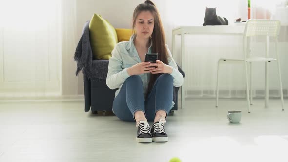 young woman sits on the floor at home and writes a message on the phone alt