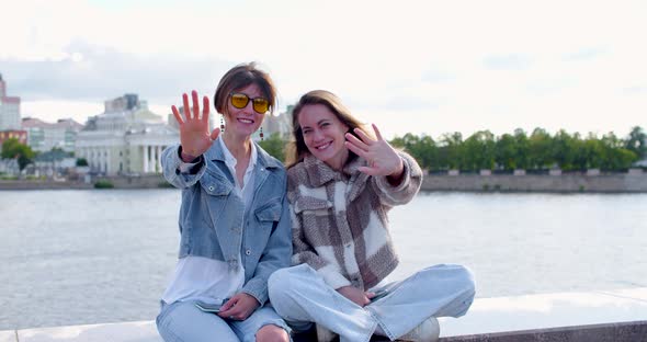 A Lesbian Couple is Sitting on the Embankment They are Waving at the Camera and Making a Heart Shape