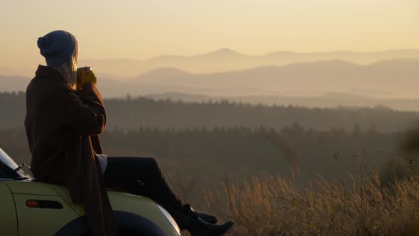 girl with mug of tea sitting on a car in beautiful mountains Sudety in November in sunset