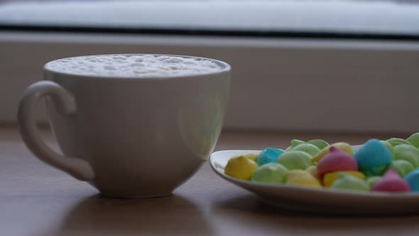 A Large Mug of Coffee with Milk or Mochacino and Colorful Sweets in a Plate on the Windowsill in