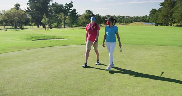 Two caucasian women playing golf wearing face masks walking together alt