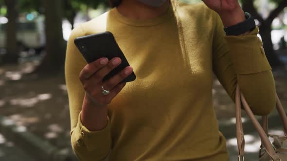 Midsection of african american woman using smartphone drinking coffee walking in city park alt