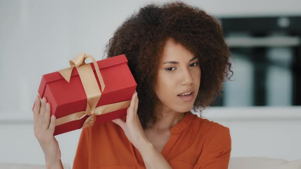Curious Interested Happy Birthday Girl African Woman with Curly Hair Shaking Wrapped Gift Box alt
