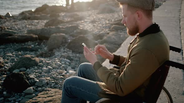 Man Sitting in Wheelchair on Quay Is Using Smartphone with Internet Swiping alt