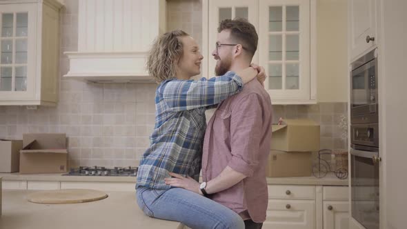 Bearded Man Stands Near Pretty Woman Sitting on Table in Big Half Empty Kitchen alt