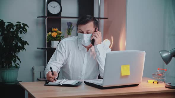 A man in a mask in front of a laptop at the workplace is talking on the phone