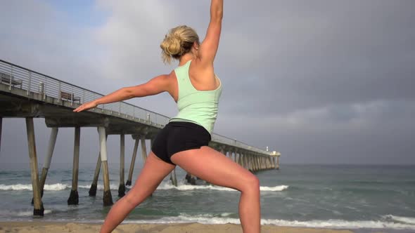 A young attractive woman doing yoga on the beach next to a pier. alt