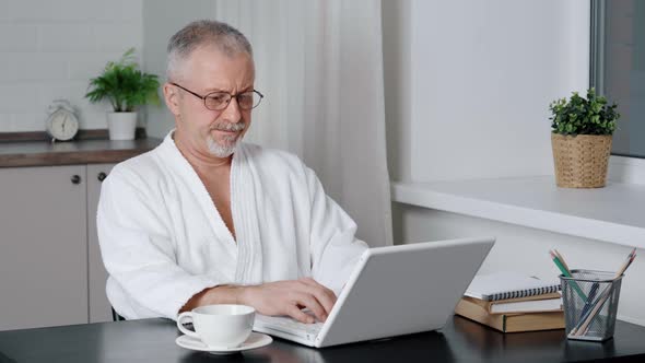 A Satisfied Mature Man Sitting at a Table in the Kitchen Looks at the Computer