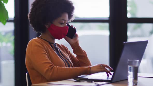 Mixed race businesswoman wearing mask sitting using a laptop in office alt