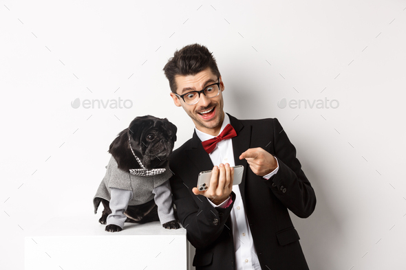 Happy dog owner showing something to pet on mobile phone, man and pug wearing fancy costumes