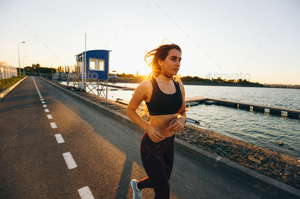Beautiful young girl with long brown hair dressed in sports clothes runs on the road along the water