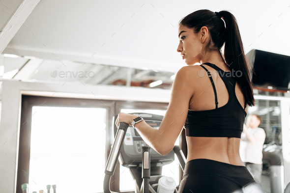Dark-haired athletic girl dressed in black sports top and tights works out on a treadmill in the gym