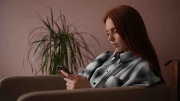Side View of Serious Redhead Young Woman Sitting on Soft Armchair and Messaging on Smartphone at alt